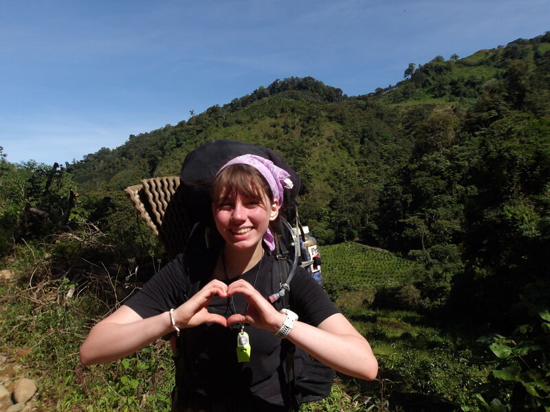 The image shows a woman with a backpack on a hiking trail. She is making a heart shape with her hands. She is smiling and appears to be enjoying the hike. The background features a lush green hillside under a blue sky.